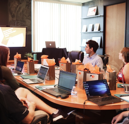 Fotografija skupine oseb na sestanku, ki sedijo za mizo z računalniki. 